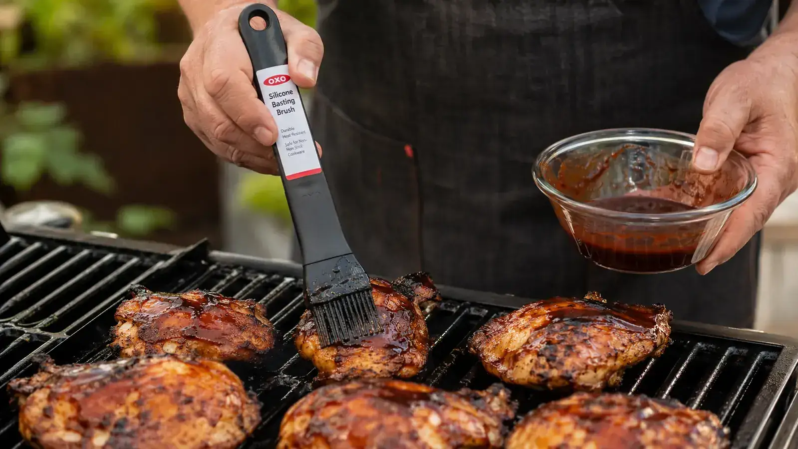 Hand using the OXO Good Grips silicone basting brush to sauce chicken thighs on a gas grill