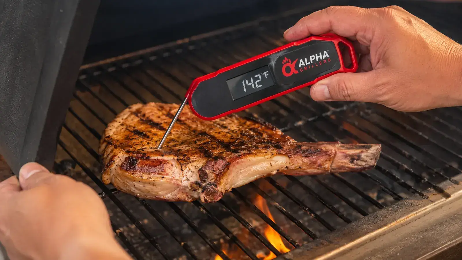 Hand inserting the Alpha Grillers thermometer probe into a thick pork chop on a charcoal grill, digital display showing 142 degrees Fahrenheit