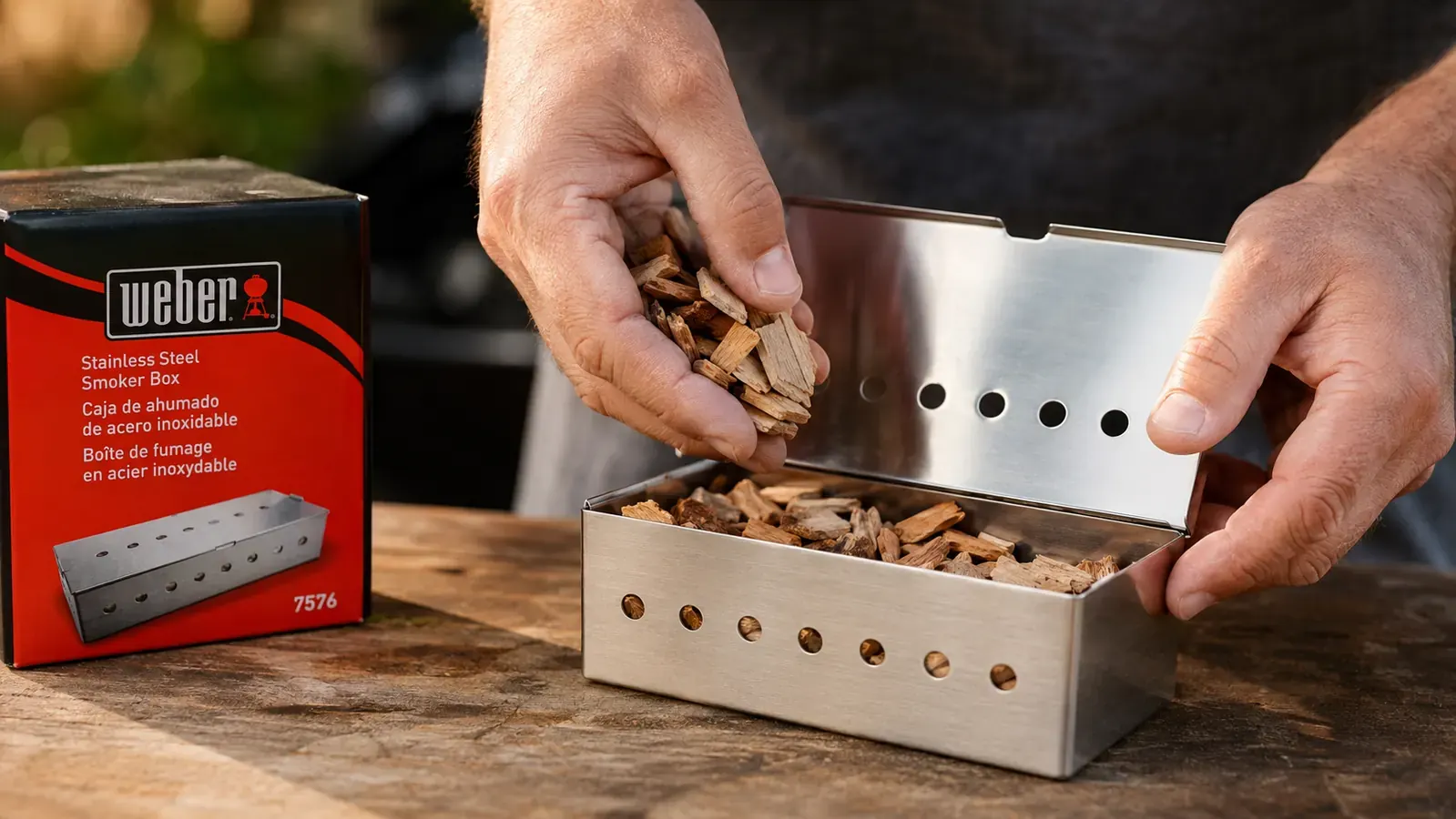 Hand filling the Weber smoker box with hickory wood chips before placing it under the grill grates