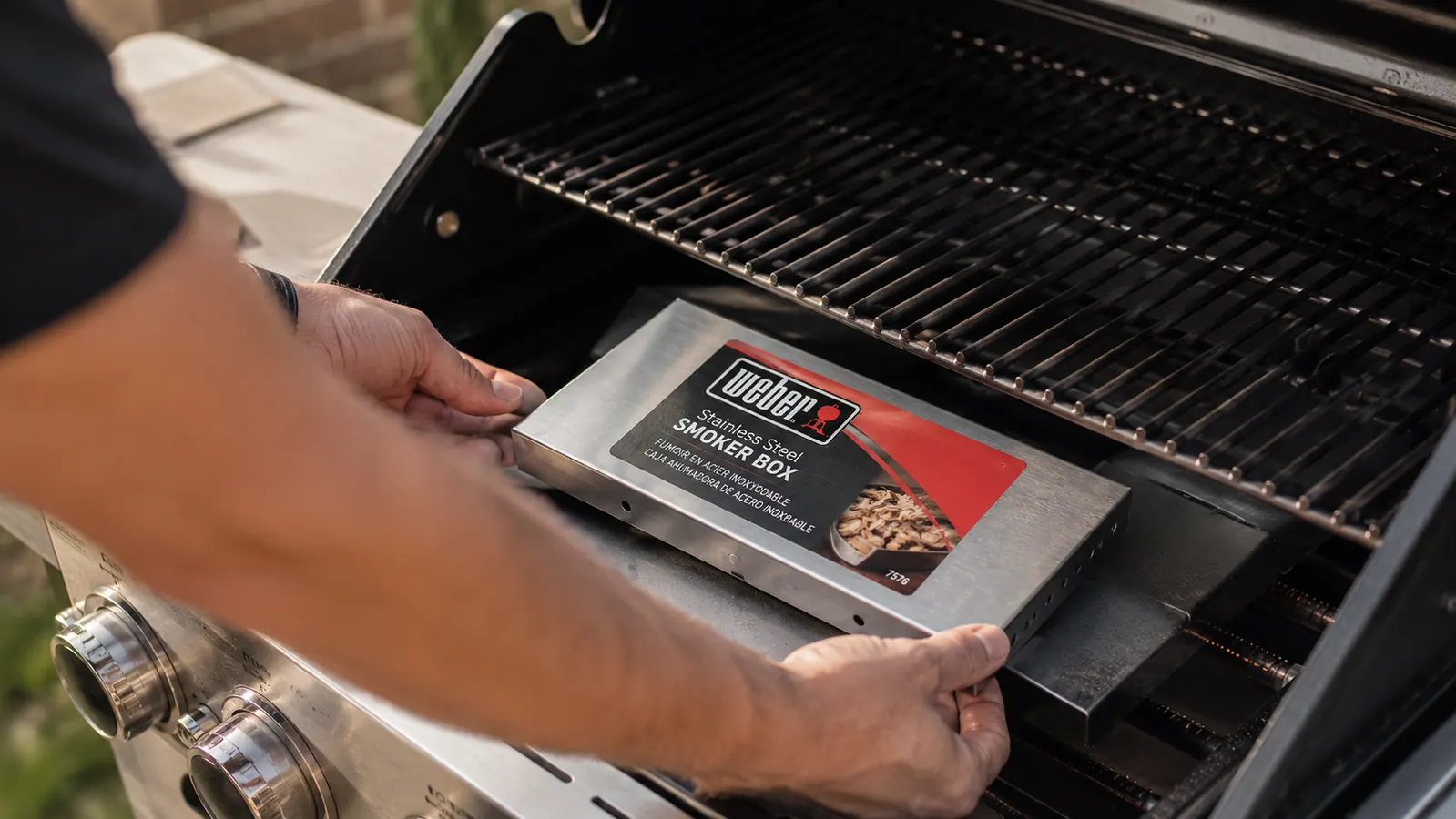 Weber smoker box placed directly on a gas grill burner cover below the cooking grates, positioned for indirect heat smoking