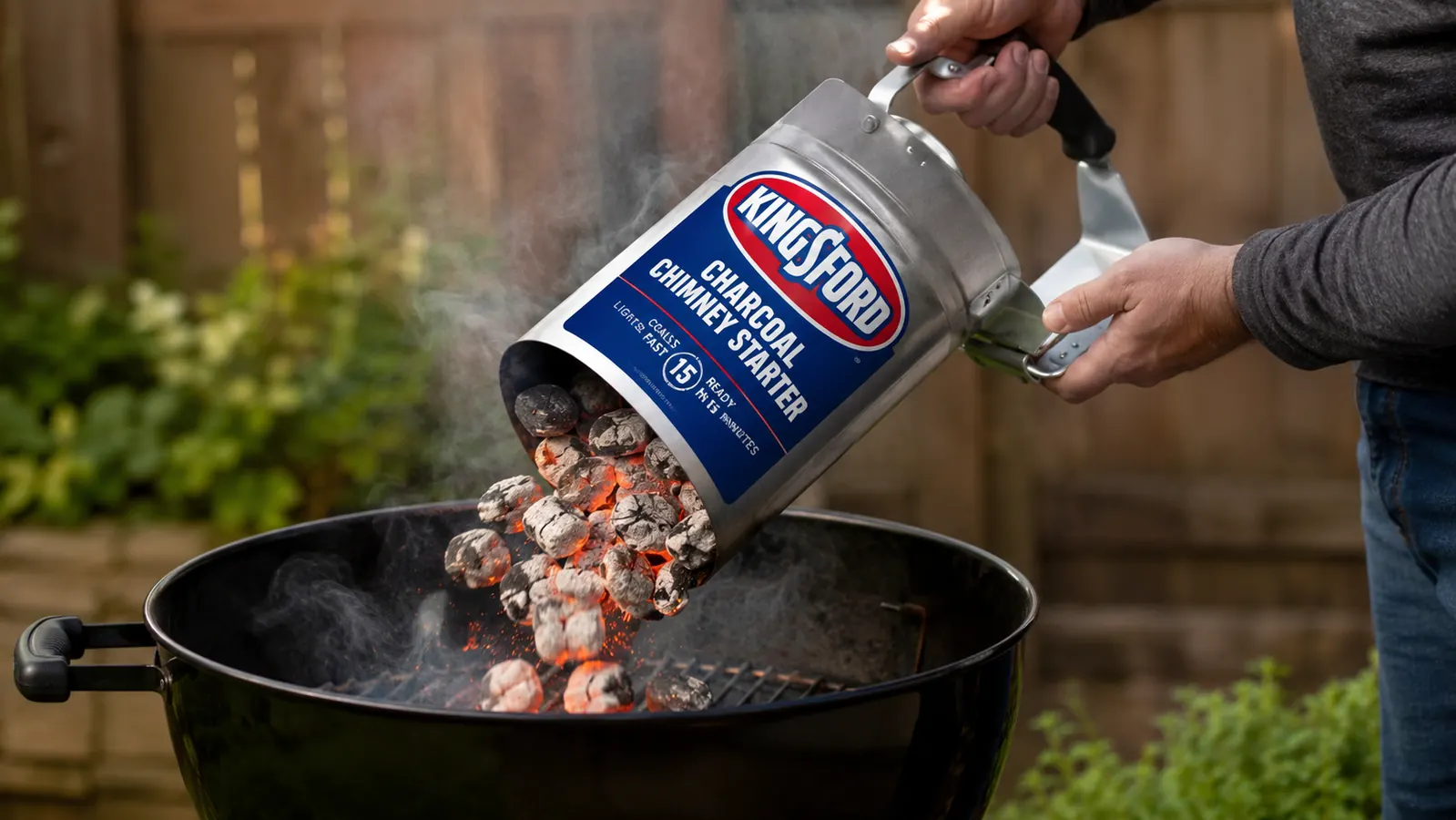 Hand holding the Kingsford chimney starter by its stay-cool handle over a charcoal grill, coals tumbling in