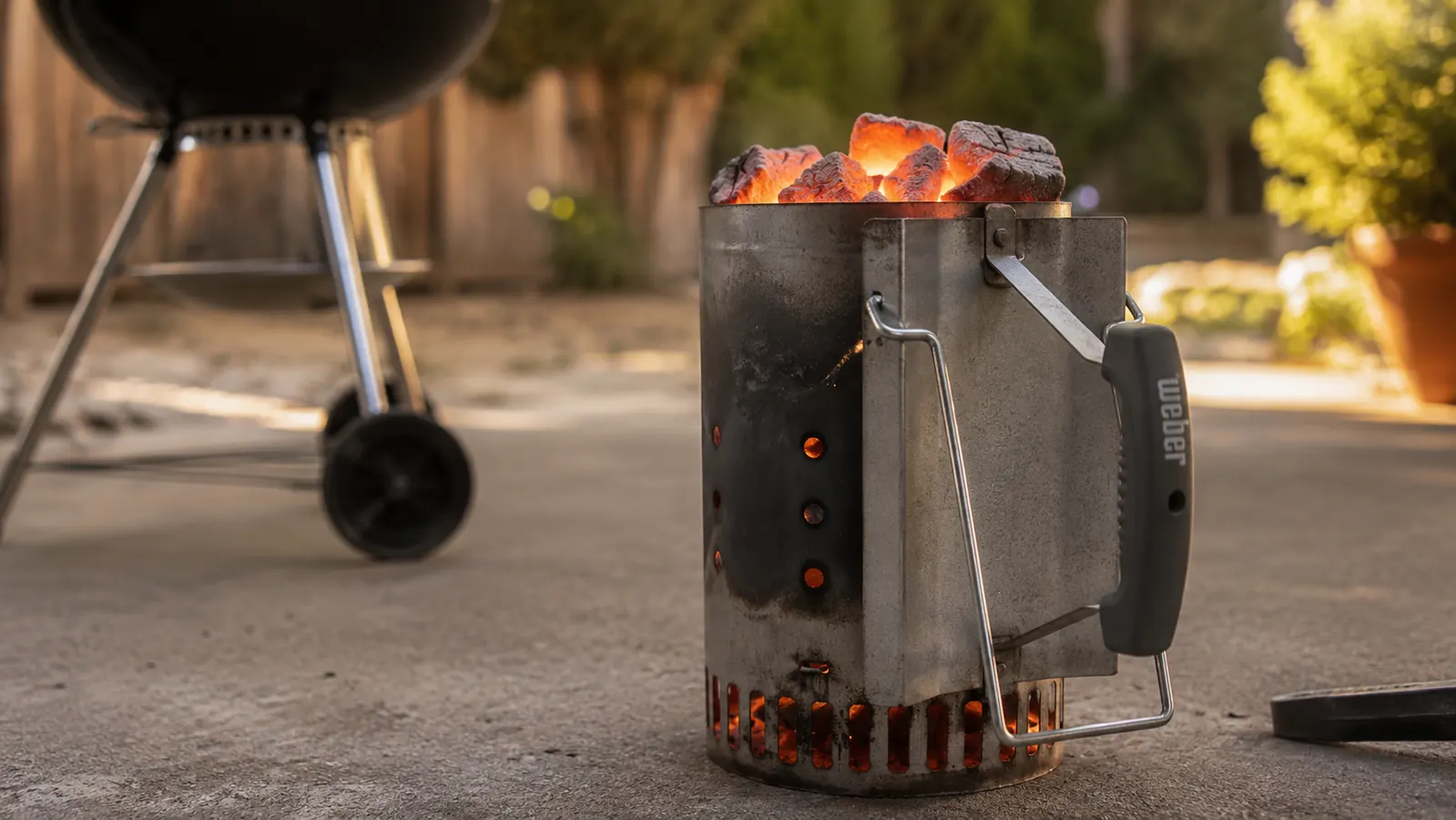 Kingsford compact chimney starter sitting on a concrete patio with orange-glowing coals visible through the vents, morning light