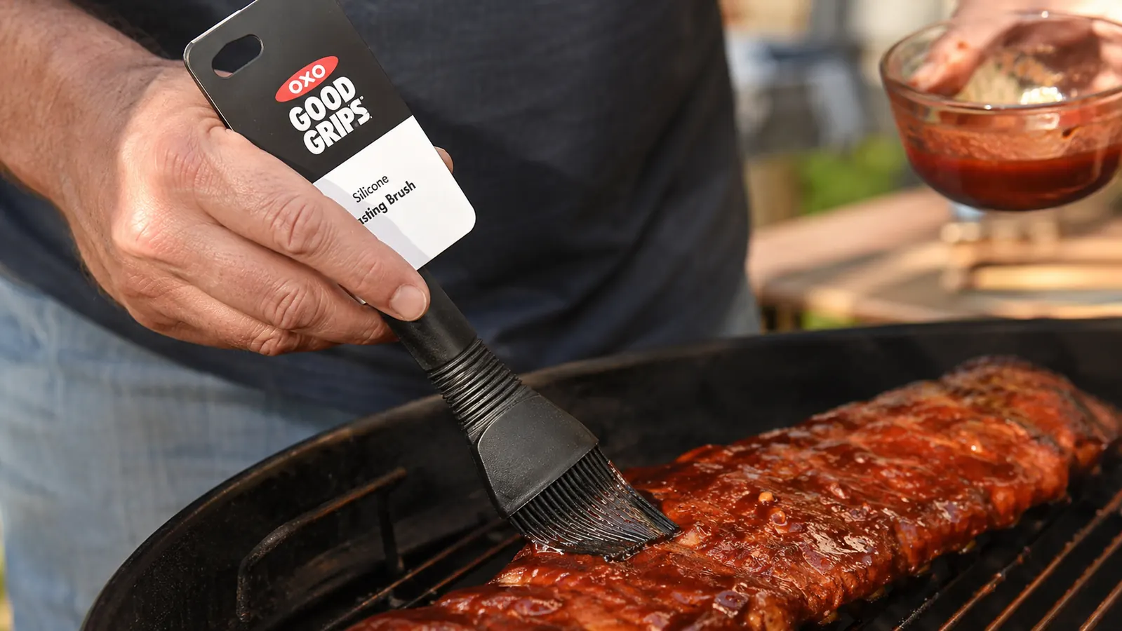 Hand holding an OXO silicone basting brush and applying sauce to pork ribs on a grill