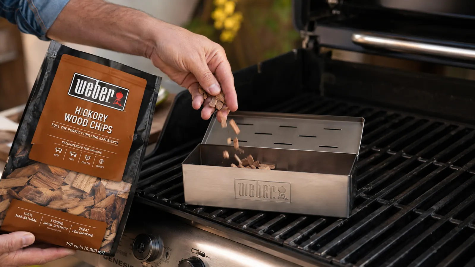 Hand loading hickory wood chips into the open Weber smoker box lid before a cook