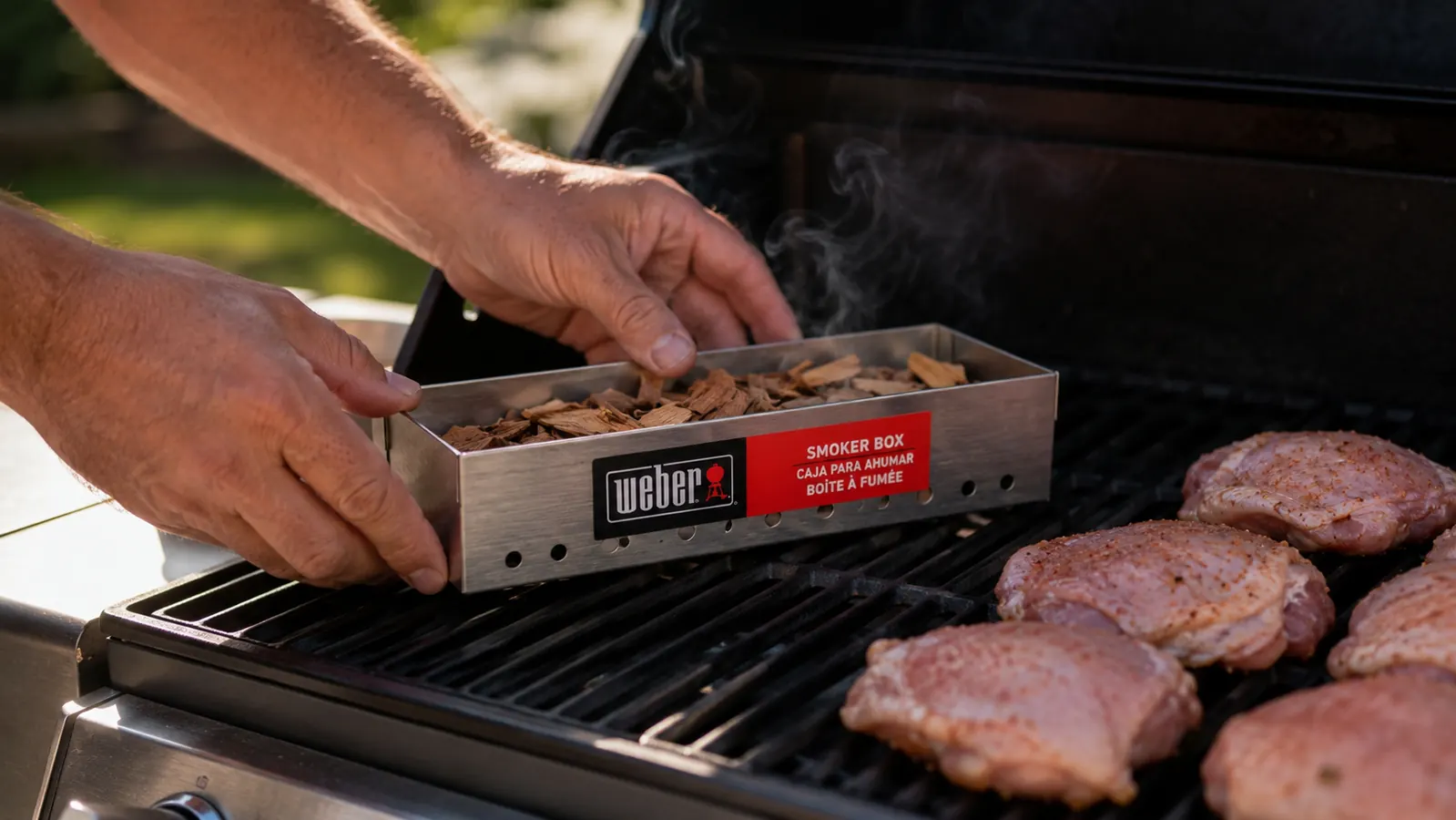 Hands placing the Weber stainless steel smoker box loaded with wood chips onto the grill grate next to chicken thighs